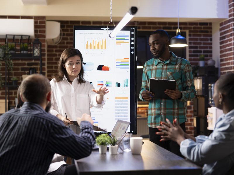 Male and female business executives stand in front of LED monitor, presenting financial data and project plans in brick wall workspace. Diverse entrepreneurs reviewing marketing trends on screen.